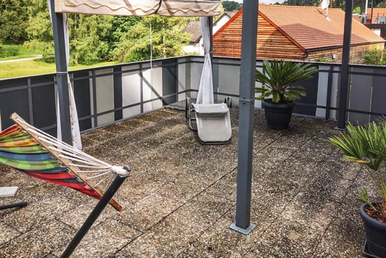 La terrazza sul tetto con vecchie piastrelle in calcestruzzo offre un’ampia area soggiorno. La superficie sporca delle piastrelle mostra evidenti segni di usura. La tenda fornisce una piacevole ombra nelle giornate calde. Vasi con palme e un’amaca creano un’atmosfera accogliente. La struttura della ringhiera in metallo e vetro completa il design moderno e aperto della terrazza.