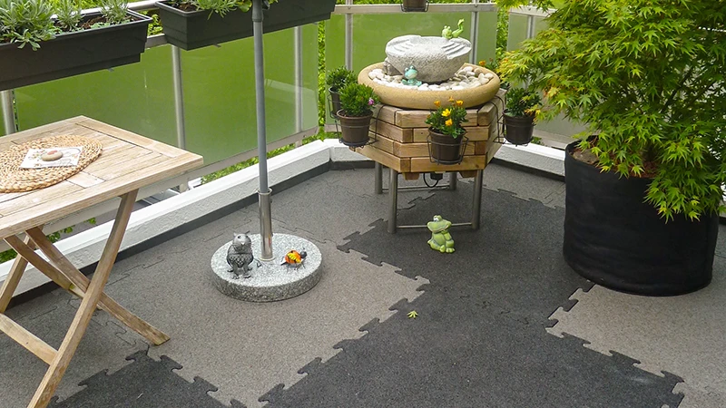Auf dem Foto ist ein einladender Balkon zu sehen, der mit zweifarbigen WARCO-Puzzleplatten ausgelegt ist. Ein kleiner Brunnen, eine Holzablage und mehrere Pflanzgefäße schaffen eine gemütliche Atmosphäre. Der Bodenbelag bietet durch seine elastische, rutschfeste Oberfläche hohen Komfort und ist zugleich witterungsbeständig. Dank der passgenauen Verlegung lassen sich Farbakzente setzen, die dem Außenbereich ein modernes Flair verleihen.