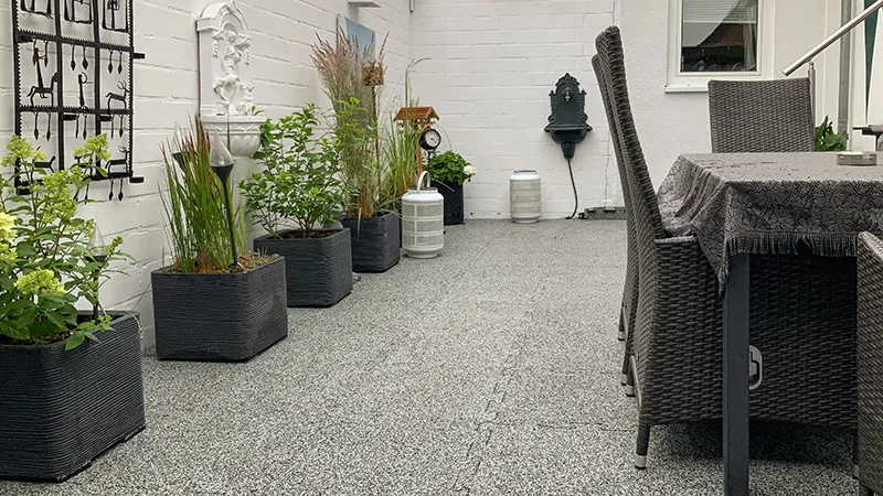 Die von einer weißen Mauer umgebene Terrasse ist wohnlich gestaltet und mit hellgrau melierten Terrassenplatten aus Gummigranulat ausgestattet. Die Platten sind durch eine Puzzleverzahnung am Rand der Platten miteinander verbunden. Aufgrund der hellgrau melierten, granitähnlichen Farbgebung ist die Verzahnung optisch kaum wahrnehmbar. So entsteht eine harmonische und gleichmäßige Terrassenfläche, die sich gut in den Innenhof einfügt.