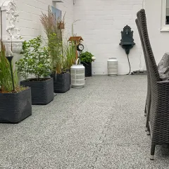 Die von einer weißen Mauer umgebene Terrasse ist wohnlich gestaltet und mit hellgrau melierten Terrassenplatten aus Gummigranulat ausgestattet. Die Platten sind durch eine Puzzleverzahnung am Rand der Platten miteinander verbunden. Aufgrund der hellgrau melierten, granitähnlichen Farbgebung ist die Verzahnung optisch kaum wahrnehmbar. So entsteht eine harmonische und gleichmäßige Terrassenfläche, die sich gut in den Innenhof einfügt.