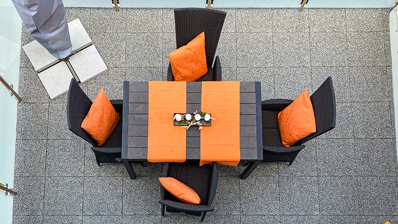 Auf dem Bild sieht man einen geräumigen Balkon, dessen Boden mit grauen WARCO-Bodenplatten ausgelegt ist. In der Mitte steht ein großer Esstisch mit schwarzen Rattanstühlen, die durch orangefarbene Kissen farbliche Akzente setzen. Der elastische und rutschfeste Bodenbelag bietet Komfort und Trittsicherheit. Zudem ist er pflegeleicht sowie witterungsbeständig und verleiht dem Außenbereich ein modernes Ambiente, das zum Verweilen einlädt.