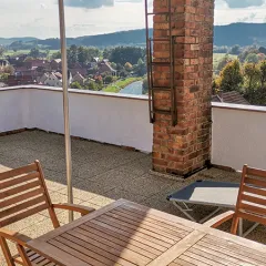Die Dachterrasse bietet einen weiten Blick über ein Dorf, einen Fluss und die umliegenden Berge. Der Boden ist mit warmen, naturfarbenen Terrassenplatten aus Gummigranulat belegt, die eine angenehme Atmosphäre schaffen. Ein Holztisch mit Stühlen und eine Liege laden zum Verweilen ein. Die gemauerte Säule und die weiße Brüstung verleihen der Dachterrasse ein harmonisches Gesamtbild.