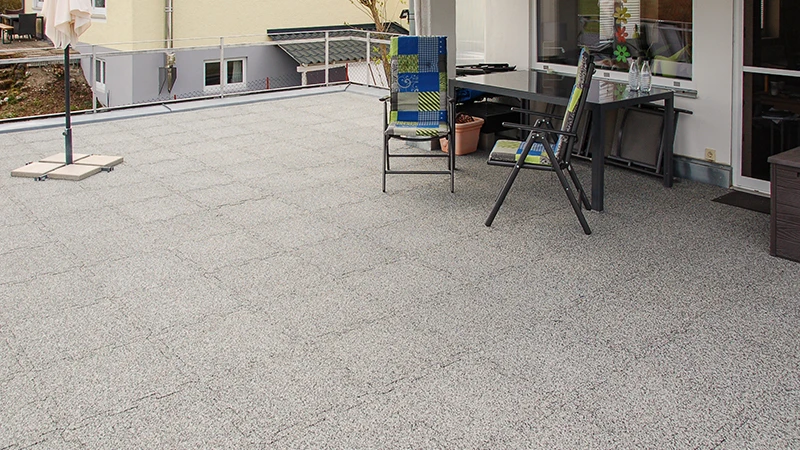 Su un terrazzo si trovano piastrelle WARCO grigie con effetto marmorizzato, realizzate in granuli di gomma legati in PU. Il loro incastro a puzzle garantisce una connessione precisa e unisce resistenza agli agenti atmosferici a una lunga durata. Il materiale, stabile nella forma, attutisce i rumori dei passi e contribuisce a un aspetto ordinato e curato. Offre inoltre una superficie antiscivolo e un elevato comfort di camminata, ideale per un utilizzo pratico.