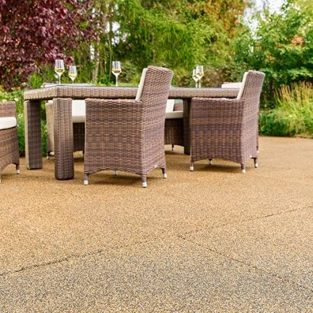 Terrazza soleggiata in giardino con piastrelle WARCO beige-marrone in granuli di gomma legati in PU. La superficie antiscivolo e permeabile all’acqua garantisce un’eccellente comodità di camminata e riduce i rumori. Un robusto tavolo da giardino con sedie intrecciate e calici di vino invita al relax. Arbusti rigogliosi e fiori variopinti creano un ambiente naturale. Questo pavimento elastico resiste al gelo, mantiene la forma e si adatta all’uso in ogni stagione.