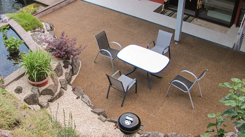 Die Terrasse aus braun-beige marmorierten Gummigranulatplatten von WARCO fügt sich harmonisch in die naturnahe Gartengestaltung ein. Der elastische Bodenbelag ist wetterfest und bietet auch barfuß ein angenehmes Laufgefühl. Der großzügige Sitzbereich mit Gartentisch und Stühlen lädt zum Verweilen ein. Der angrenzende Teich mit Natursteinen und üppiger Bepflanzung schafft eine idyllische Atmosphäre. Der runde Grill ergänzt die gemütliche Outdoor-Oase.
