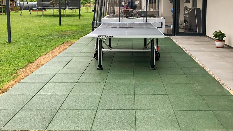 Die grüne Terrasse mit quadratischen Gummigranulatplatten von WARCO bietet eine robuste und rutschfeste Bodenfläche für Spiel und Sport im Garten. Der elastische Bodenbelag dämpft Stöße und sorgt für ein angenehmes Laufgefühl, ideal für sportliche Aktivitäten wie Tischtennis. Die harmonische Farbgebung fügt sich nahtlos in die natürliche Umgebung ein. Im Hintergrund sorgen ein Trampolin und der angrenzende Wintergarten für zusätzliche Freizeitmöglichkeiten.