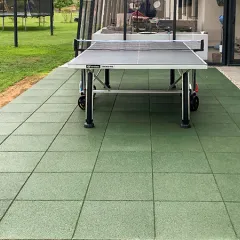 Die grüne Terrasse mit quadratischen Gummigranulatplatten von WARCO bietet eine robuste und rutschfeste Bodenfläche für Spiel und Sport im Garten. Der elastische Bodenbelag dämpft Stöße und sorgt für ein angenehmes Laufgefühl, ideal für sportliche Aktivitäten wie Tischtennis. Die harmonische Farbgebung fügt sich nahtlos in die natürliche Umgebung ein. Im Hintergrund sorgen ein Trampolin und der angrenzende Wintergarten für zusätzliche Freizeitmöglichkeiten.