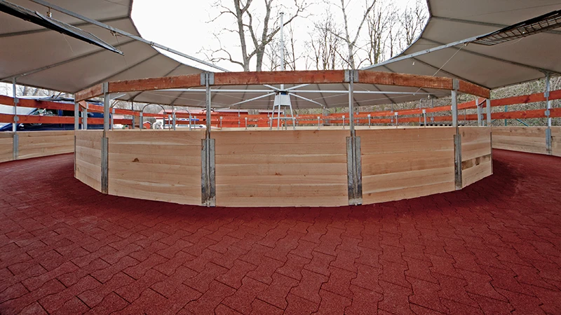 La foto mostra una moderna giostra coperta per cavalli. Il percorso è completamente rivestito con mattonelle in gomma rossa – antiscivolo, drenanti e delicate sulle articolazioni. La superficie elastica consente movimenti sicuri in ogni condizione climatica e andatura. Il fondo è stabile, durevole e richiede pochissima manutenzione. La struttura è ideale per centri ippici, allevamenti e cliniche veterinarie equine che puntano su superfici adatte agli animali, resistenti e a bassa manutenzione.
