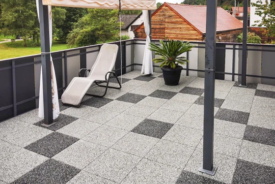 La terrazza sul tetto ristrutturata è pavimentata con piastrelle effetto granito disposte in un motivo geometrico di quadrati grigio chiaro e grigio scuro, creando un'atmosfera moderna ed elegante. Il pavimento robusto in gomma garantisce sicurezza antiscivolo e comfort. Una tenda parasole offre ombra nelle giornate calde. Un vaso con palma e una sdraio completano il design accogliente della terrazza. La struttura in metallo e vetro del parapetto dona alla terrazza un aspetto aperto e arioso.
