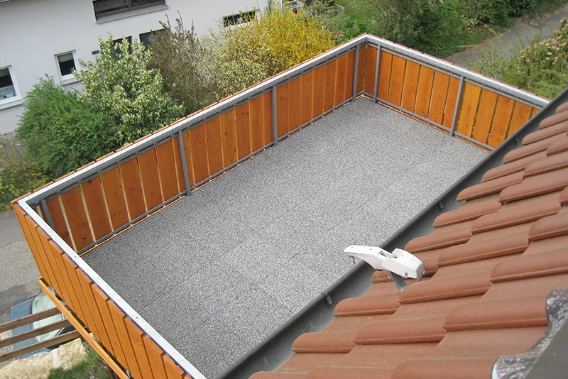 Hellgraue Balkonplatten auf einem kleinen Balkon mit Holzgeländer. Graue Balkonplatten liegen auf mit Hozlgeländer gesichertem Balkon. Der Blick fällt vom Dach in den Hof, in dem Büsche und Bäume wachsen.