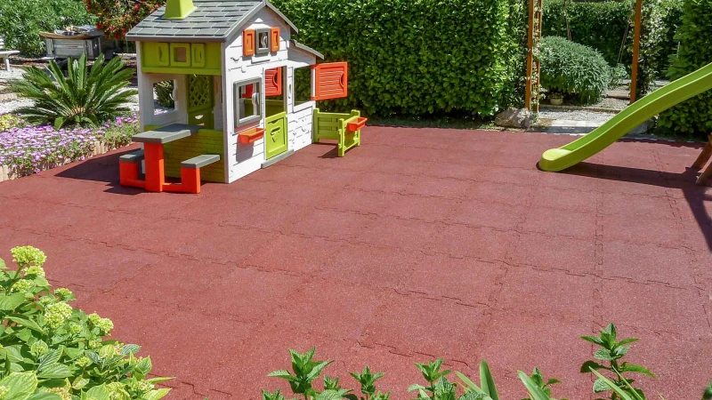 Il parco giochi con la casetta nel giardino privato. Il parco giochi per bambini in giardino. La casetta di plastica è stata installata su un'ampia superficie di lastre ammortizzanti WARCO in colore rosso.