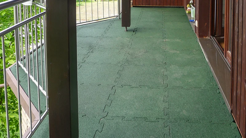 Auf dem Foto ist ein langgezogener Balkon zu sehen, der mit grünen WARCO-Bodenplatten in Puzzle-Form ausgelegt ist. Die rutschfeste und elastische Oberfläche bietet Sicherheit und Komfort, auch bei feuchtem Wetter. Dank der passgenauen Verlegung entsteht eine langlebige, pflegeleichte Lösung, die den Untergrund schützt. So wird der Außenbereich zu einem angenehmen Ort zum Verweilen und Entspannen.