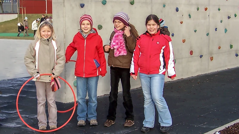 Kinder spielen auf Outdoor-Fallschutzmatten neben einer Kletterwand im Freien.