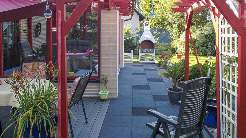 Die erweiterte Terrasse am Haus bietet mit den quadratischen Gummigranulatplatten von WARCO in mittelgrau und dunkelgrau eine elegante und wetterfeste Bodenfläche. Die marmorierte Optik der Platten verleiht der Terrasse einen modernen und ansprechenden Charakter. Dank der rutschfesten Oberfläche ist die Nutzung bei jedem Wetter sicher und komfortabel. Der einladende Sitzbereich wird durch Pflanzen und dekorative Elemente ergänzt. Im Hintergrund sorgt der Grillkamin für eine gemütliche Atmosphäre und lädt zu entspannten Stunden im Freien ein.