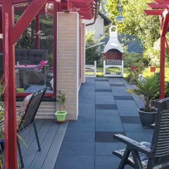 Die erweiterte Terrasse am Haus bietet mit den quadratischen Gummigranulatplatten von WARCO in mittelgrau und dunkelgrau eine elegante und wetterfeste Bodenfläche. Die marmorierte Optik der Platten verleiht der Terrasse einen modernen und ansprechenden Charakter. Dank der rutschfesten Oberfläche ist die Nutzung bei jedem Wetter sicher und komfortabel. Der einladende Sitzbereich wird durch Pflanzen und dekorative Elemente ergänzt. Im Hintergrund sorgt der Grillkamin für eine gemütliche Atmosphäre und lädt zu entspannten Stunden im Freien ein.