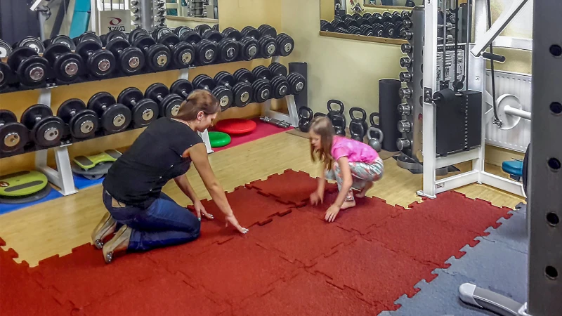 Una madre e una figlia posano dei tappeti sportivi rossi, in granuli di gomma, per proteggere il pavimento sul parquet della palestra di casa.