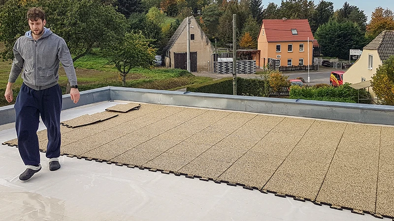 Auf dem Foto ist ein weitläufiger Balkon zu sehen, auf dem eine Person beigen WARCO-Puzzleplatten verlegt. Durch das präzise Puzzlesystem lassen sich die Bodenplatten rasch zusammenfügen. Die rutschfeste, witterungsbeständige Oberfläche schützt den Untergrund und bietet eine begehbare Fläche für viele Nutzungsmöglichkeiten. So entsteht ein sicherer, komfortabler und pflegeleichter Außenbereich, der zum Entspannen einlädt. Zudem sorgt die Dämmwirkung für eine angenehme Trittschalldämpfung.