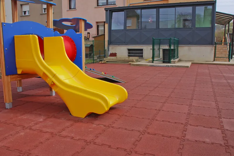 Spielplatz mit WARCO Fallschutzmatten aus Gummi in Ziegelrot Das Bild zeigt einen Spielplatz mit einem gelben Doppelrutschenturm im Vordergrund, der auf einer Fläche aus WARCO Fallschutzmatten aus Gummi steht. Die Matten sind ziegelrot und durch die Schwalbenschwanzverzahnung an den Plattenrändern formschlüssig zu einem stabilen Plattenteppich verbunden. Im Hintergrund sieht man ein Gebäude. Der Bodenbelag bietet eine stoßdämpfende Oberfläche, die das Verletzungsrisiko bei Stürzen minimiert und gleichzeitig eine langlebige und witterungsbeständige Lösung für den Außenbereich darstellt.