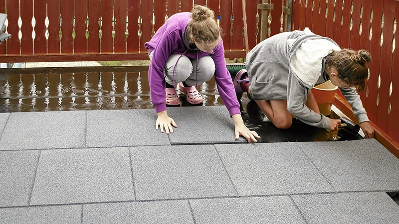 Auf dem Foto verlegen zwei Personen graue WARCO-Bodenplatten auf einer nassen Holzterrasse. Dank des passgenauen Formats lassen sich die Platten schnell und einfach aneinanderfügen. Der rutschfeste und wasserdurchlässige Bodenbelag ist optimal für Außenbereiche geeignet. Die robusten Platten schützen den Untergrund und sorgen für einen sicheren, bequemen Laufkomfort. So entsteht im Handumdrehen eine strapazierfähige und pflegeleichte Terrasse.