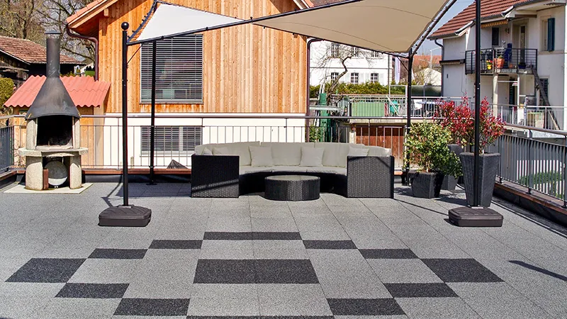 Eine stilvoll gestaltete Dachterrasse, ausgelegt mit Terrassenplatten aus PU-gebundenem Gummigranulat von WARCO in mittlerem Grau und Dunkelgrau. Die Platten bilden ein ansprechendes, geometrisches Muster und schaffen eine robuste, witterungsbeständige Bodenfläche. Ein großzügiges Sonnensegel spendet Schatten für die Lounge-Garnitur mit Sitzbank und rundem Tisch. Ein Grillkamin aus Stein bietet eine gemütliche Atmosphäre für entspannte Stunden im Freien.