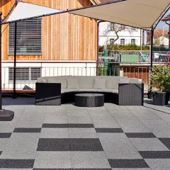 Eine stilvoll gestaltete Dachterrasse, ausgelegt mit Terrassenplatten aus PU-gebundenem Gummigranulat von WARCO in mittlerem Grau und Dunkelgrau. Die Platten bilden ein ansprechendes, geometrisches Muster und schaffen eine robuste, witterungsbeständige Bodenfläche. Ein großzügiges Sonnensegel spendet Schatten für die Lounge-Garnitur mit Sitzbank und rundem Tisch. Ein Grillkamin aus Stein bietet eine gemütliche Atmosphäre für entspannte Stunden im Freien.