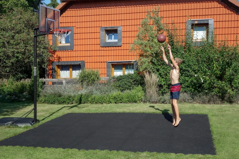 Il piccolo campo da basket nel giardino realizzato con le lastre per giocare a palla di WARCO in colore Antracite è stato creato dalla famiglia. Un ragazzo gioca a basket.