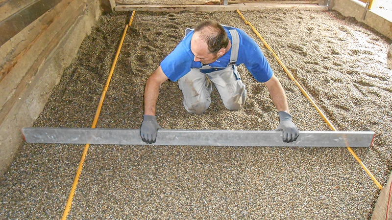 Installazione di un nuovo fondo in una scuderia: Un uomo livella uno strato di ghiaia con una staggia. La ghiaia funge da letto di posa per un pavimento da box in granuli di gomma. L’area è delimitata lateralmente da pareti in legno o cemento. Il lavoro viene eseguito con cura e precisione – tipico di una corretta preparazione di un fondo stabile adatto ai cavalli.