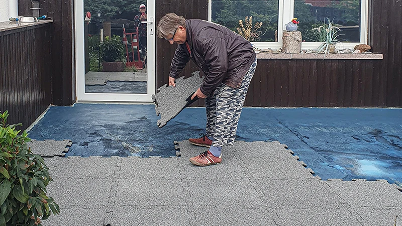 Una terrazza è in fase di ristrutturazione. Una signora anziana posa la successiva piastrella da 18 mm di spessore con incastro sulla terrazza già parzialmente coperta con nuove piastrelle. Un foglio impermeabilizzante funge da base per il nuovo pavimento in piastrelle di gomma. L’incastro preciso delle piastrelle consente una posa facile e stabile.