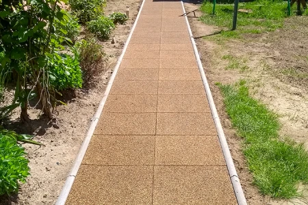 Un vialetto da giardino dritto in piastrelle quadrate di gomma con cordolo laterale attraversa l’aiuola. Le piastrelle marroni in granuli di gomma offrono una superficie antiscivolo, resistente alle intemperie e facile da mantenere. Il vialetto garantisce un passaggio sicuro e pulito in ogni condizione. Ideale per giardini, cortili e accessi.
