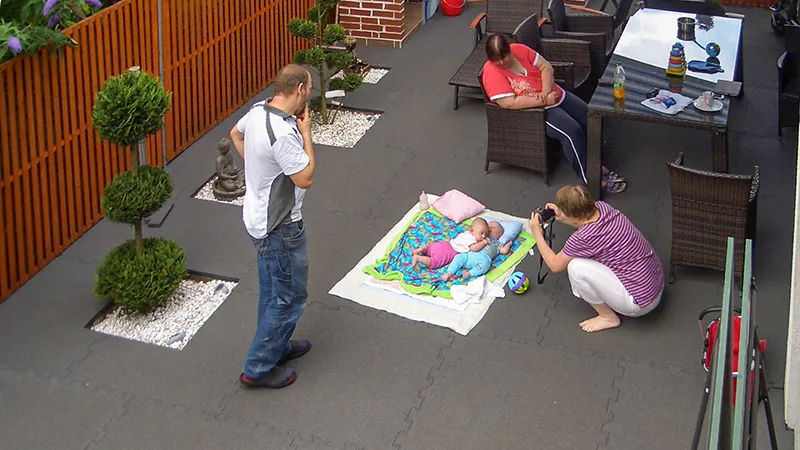 Auf der familienfreundlich gestalteten Terrasse liegen großformatige, graue Klickfliesen aus Gummigranulat. Die Platten sind durch eine Puzzleverzahnung am Rand miteinander verbunden. Zwei Babys liegen auf einer Decke, während eine Frau sie fotografiert. Zwei weitere Erwachsene stehen bzw. sitzen entspannt daneben. Die Terrasse bietet eine wohnliche Atmosphäre und viel Platz für gemeinsame Aktivitäten im Freien.