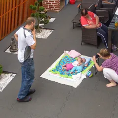 Auf der familienfreundlich gestalteten Terrasse liegen großformatige, graue Klickfliesen aus Gummigranulat. Die Platten sind durch eine Puzzleverzahnung am Rand miteinander verbunden. Zwei Babys liegen auf einer Decke, während eine Frau sie fotografiert. Zwei weitere Erwachsene stehen bzw. sitzen entspannt daneben. Die Terrasse bietet eine wohnliche Atmosphäre und viel Platz für gemeinsame Aktivitäten im Freien.