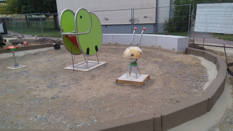 Ein Kleinkinderspielplatz in Form einer runden Insel befindet sich im Bau. Die Spielgeräte sind bereits aufgestellt, die Tragschicht für den Boden ist eingebaut. Die Fläche wird mit Kettenelementen, d.h. miteinander verbundenen Blockstufen aus Gummigranulat, eingefasst.