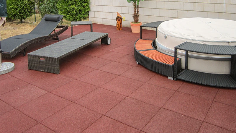 Un’ampia area esterna con piastrelle da terrazza rosse WARCO in granuli di gomma legati in PU, che offrono una calpestabilità confortevole e un’elevata resistenza. Arredata con comodi mobili da giardino e una vasca idromassaggio che si integra perfettamente nell’insieme. La superficie elastica attutisce i passi, resiste alle intemperie e richiede poca manutenzione. Grazie alla sua finitura antiscivolo, rappresenta una soluzione ideale per rilassarsi con stile all’aperto. Perfetta per terrazze moderne che uniscono funzionalità e comfort.