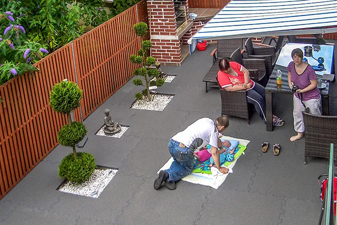Eine Familie genießt ihre freie Zeit auf einer als Wohlfühl-Oase gestalteten Terrasse mit grauen Terrassenbelag von WARCO. Das Baby liegt auf eine Kinderdecke auf den Terrassenplatten Eine junge Familie hat sich ihre Dachterrasse als Wohlfühloase mit Sitzgruppe, Grill, Bonsai und dämmendem Terrassenbelag von WARCO im Farbdesign betongrau gestaltet. Das Baby der Familie liegt, nur durch eine Kinderdecke getrennt, direkt auf dem elastischen Terrassenbelag.